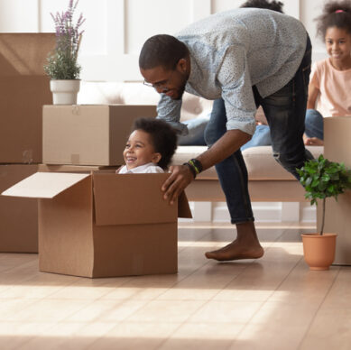 Happy young african American father playing together with preschooler son ride sitting in cardboard box, excited smiling black dad feel playful have fun with cute little kid entertain on moving day